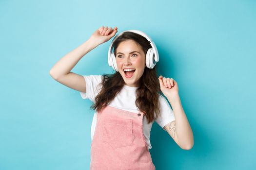 Image of cheerful young pretty girl dancing in headphones, listening music in app and jumping excited, standing over blue background.