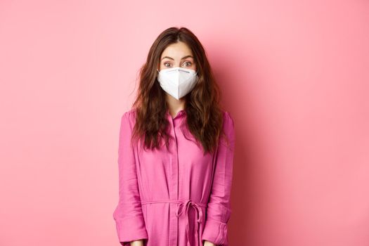 Covid-19, pandemic and lifestyle concept. Excited young woman wearing respirator while going out, social distance herself during quarantine, pink background.