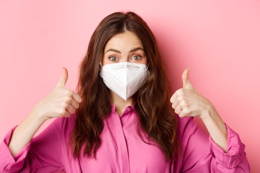 Coronavirus, preventive measures and health concept. Happy beautiful lady in medical respirator from covid-19, shows thumbs up in approval, praise good choice, pink background. Copy space
