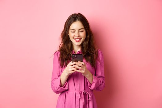 Young woman shopping, chatting on mobile phone, smiling while reading smartphone screen, standing against pink background. Copy space