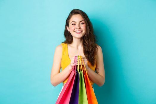 Image of cheerful girl going shopping, holding bags with purchases and smiling at camera, standing against blue background.