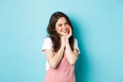Portrait of silly and cute young woman giggle flirty, smiling and looking coquettish at camera, blushing, having summer outfit and bright make up, standing over blue background.