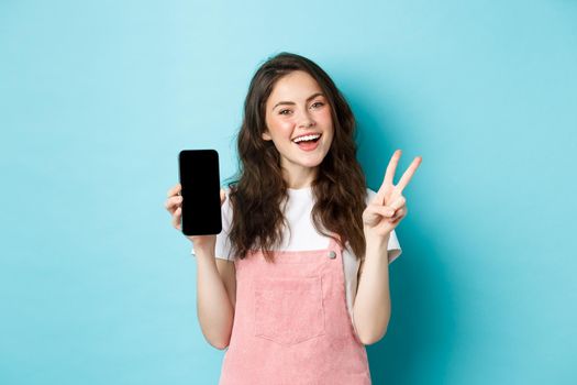 Cute young woman smiling and showing v-sign with empty smartphone screen, demonstrate app or mobile store, standing against blue background.