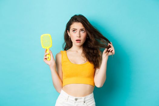 Young woman worried about her hair condition, showing brush and looking at upper left corner with concerned face, standing over blue background.