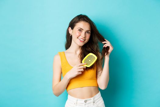 Beauty. Attractive young woman brushing her hair with hairbrush and looking dreamy at top copy space, smiling, standing against blue background.