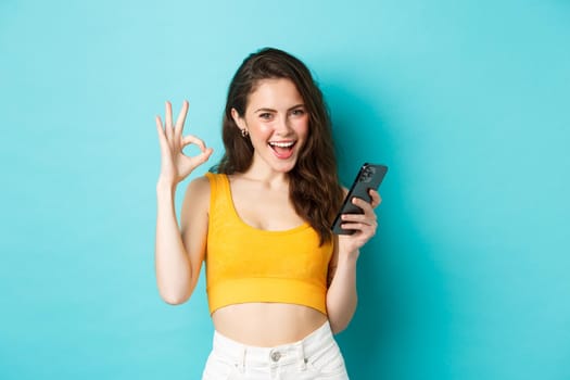Technology and lifestyle concept. Confident attractive woman say yes, showing okay sign to something online, holding smartphone, standing over blue background.