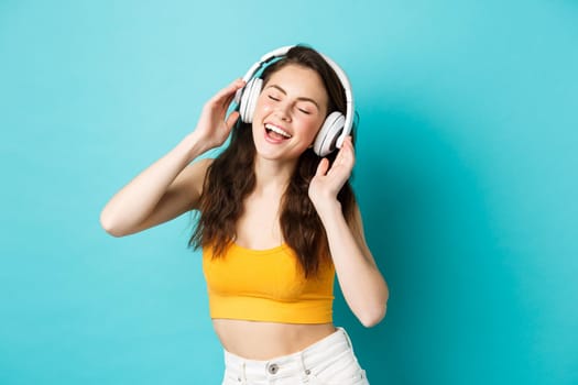 Young woman in summer clothes listening music, wearing headphones and singing along favorite song, dancing in earphones, standing against blue background.