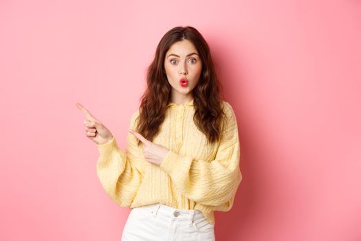 Surprised brunette woman showing promotional text, gasping amused, pointing fingers aside to left promo, standing against pink background.