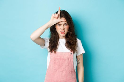 Sassy young woman despise her rival, showing loser gesture on forehead, making letter L with hand and staring with dismay at camera, standing against blue background.