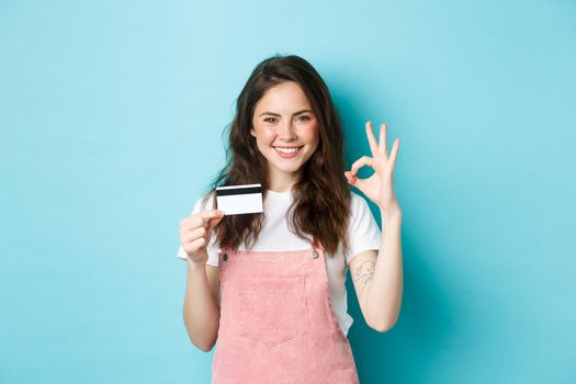 Beautiful sassy girl recommending plastic credit card, showing okay sign and looking confident at camera, smiling satisfied, standing over blue background.