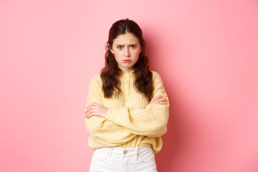 Cute silly girl feeling offended and sad, trying to comfort herself with self hug, frowning and looking at camera insulted, about to cry, standing against pink background.