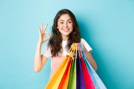 Image of happy young glamour woman shop in stores with discounts, showing okay sign, holding shopping bags, looking excited at camera, blue background.