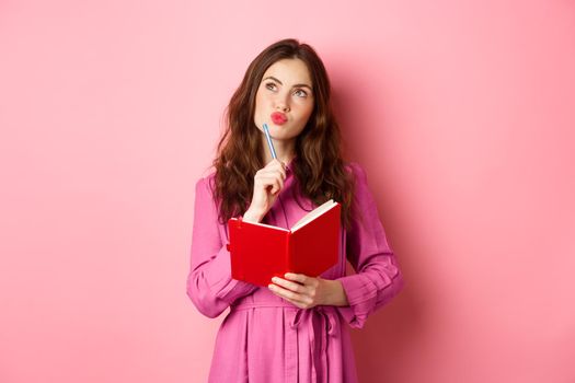 Beautiful young woman look thoughtful, writing in notebook, holding planner or diary, plan her schedule, standing against pink background.