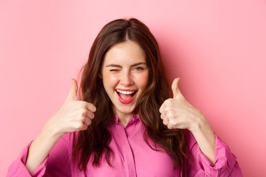 Cheeky glamour girl winking at you and shows thumbs up, praise great work, good choice, standing over pink background.