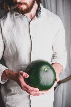 Bearded man in linen shirt holds in his hands whole watermelon