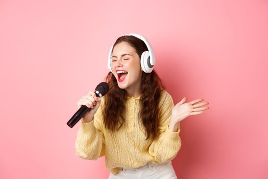 Portrait of young woman singing karaoke, wearing headphones and performing song, holding microphone, standing against pink background.