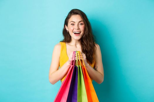 Young pretty woman holding shopping bags, smiling excited at camera, buying with discounts, standing over blue background.