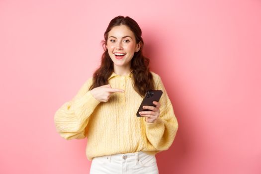 Young attractive woman holding smartphone, pointing at screen, promoting, talking about her social media page, standing over pink background.
