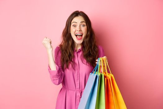 Excited girl scream of joy, making fist pump, holding shopping bag and rejoicing, standing in dress over pink background.