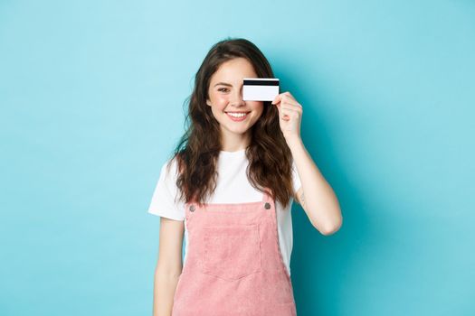 Cute young woman showing plastic credit card on eye and smiling, shopping with it, standing over blue background.
