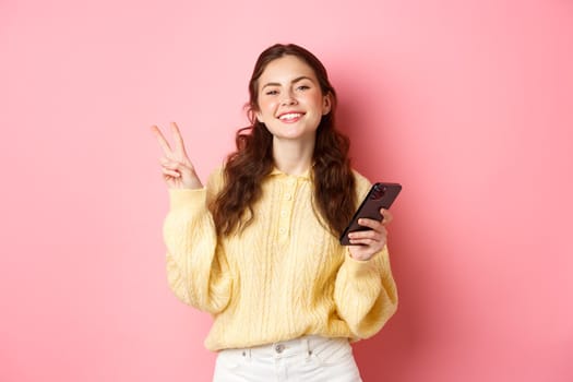 Technology and online shopping. Happy young woman smiling pleased, holding smartphone, showing v-sign and look positive at camera, standing over pink background.
