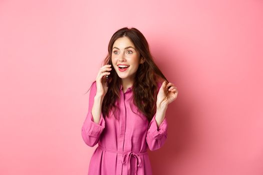 Glamour girl talking on phone and playing with hair, looking happy and relaxed during conversation, calling friend, standing against pink background.