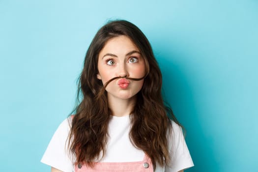 Close up portrait of funny silly girl playing, making squinting face and moustache with hair strand above lips, standing over blue background.