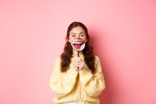 Young smiling woman showing her perfect whitened teeth smile with magnifying glass, promo of dental clinic, stomatology and healthcare concept, standing against pink background.