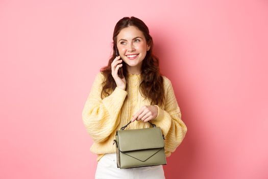 Beautiful and stylish lady having phone call, smiling while talking with someone on cellphone, holding purse, standing against pink background.