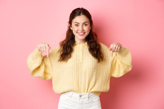 Portrait of young attractive female model pointing down at copy space, showing promotional text or logo, standing against pink background.