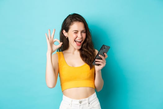 Technology and lifestyle concept. Cheerful brunette female model say yes, winking and showing okay sign, holding mobile phone, standing over blue background.