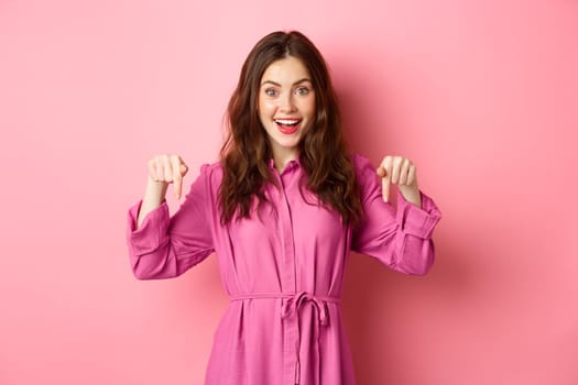 Excited smiling woman in stylish clothes pointing fingers down, looking happy and amused, checking out promo offer, standing against pink background.