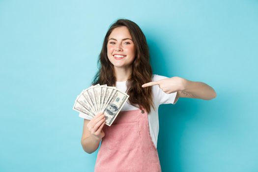 Portrait of cute girl smiling proud and satisfied, pointing finger at money dollar, showing dollar bills, standing over blue background.