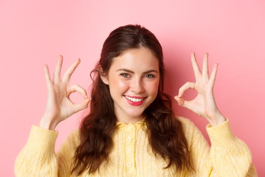 Close up of young determined woman praise good choice, showing thumbs up in approval, say yes, recommend and guarantee quality, standing over pink background.