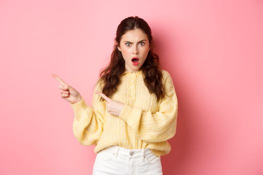 Shocked and frustrated young brunette woman, gasping and staring startled at camera, pointing fingers left at copy space, standing against pink background.