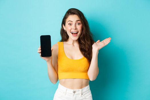 Attractive young woman in glamour summer clothes, showing empty smatphone screen, gasping amazed, recommending app, standing against blue background.