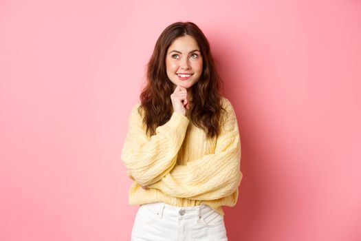 Hmm very interesting. Thoughtful young beauty girl looking aside at logo, reading promotional text and thinking, smiling pleased, pink background.