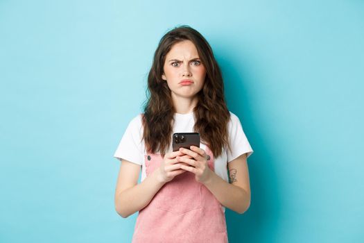 Disappointed girl frowning and grimacing displeased after using smartphone app, holding phone and express dislike, standing over blue background.