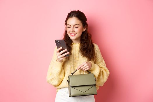 Glamour girl holding purse and texting message on smartphone, reading mobile phone screen with carefree smile, standing over pink background.