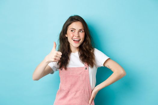 Smiling cute teen girl with beautiful blush and glamour make up, showing thumb up in approval, look at upper left corner banner, recommending store, standing against blue background.