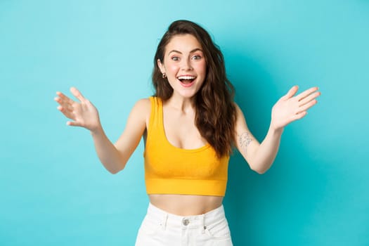 Summer and lifestyle concept. Beautiful woman look surprised and happy to see you, spread hands sideways and smiling happy, greeting person, standing over blue background.