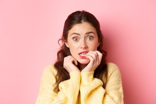 Close up of worried silly girl scared of something, biting fingers and looking nervous at camera, standing against pink background. Copy space