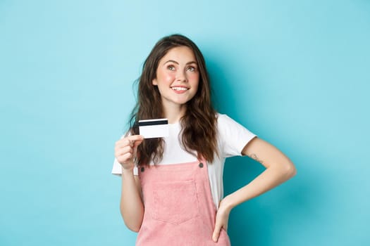 Thoughtful girl thinking of shopping, holding plastic credit card and look up with pensive face, making choice what to buy, standing over blue background.