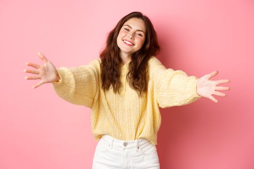 Young beautiful woman reaching hands forward to hug you, smiling friendly, inviting for free cuddles, standing against pink background.