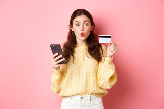 Technology and online shopping. Excited girl shopper showing plastic credit card and holding mobile phone, paying with smartphone, order online, standing against pink background.