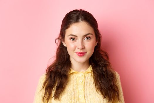 Close up of young 20s female model with cute feminine make up, smiling and looking hopeful at camera, standing against pink background.