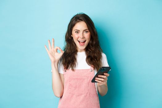 Image of excited and pleased young woman say yes after using smartphone app, online shopping with mobile phone and showing OK sign, standing over blue background.