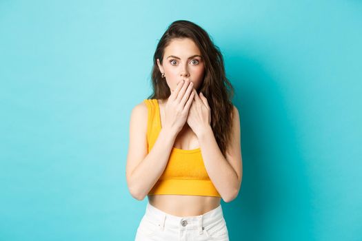 Portrait of attractive modern girl looks shocked after hear gossips, cover mouth with hands and gasping, staring with amazement at camera, standing over blue background.