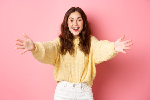 Portrait of beautiful friendly girl stretch out hands and smiling, reaching towards camera to give hug, making warm welcome, standing against pink background.