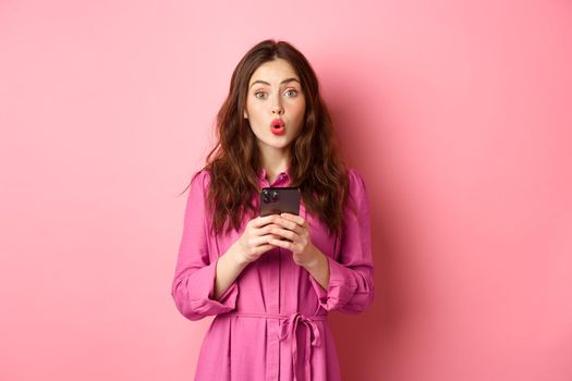 Image of surprised young woman gasping and saying wow, staring impressed at camera after reading news online on mobile phone, standing with smartphone against pink background. Copy space